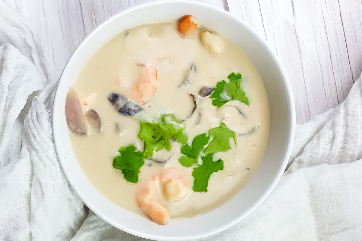 A bowl of creamy whole30 Thai coconut soup with shrimp, mushrooms, and fresh cilantro garnish sits on a white surface with a white cloth nearby.