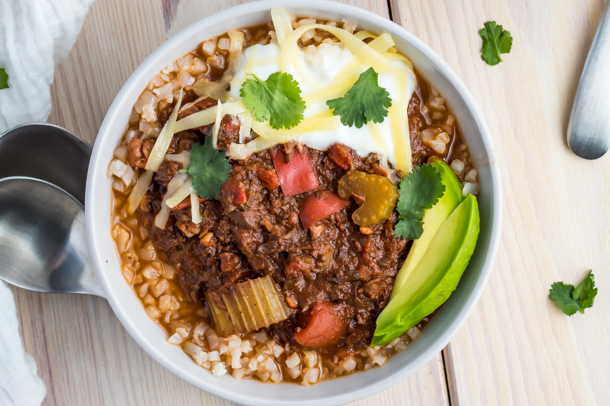 A bowl of whole30 vegan chili topped with shredded cheese, sour cream, cilantro, and avocado slices sits beside two spoons on a light wooden surface.