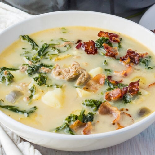 A bowl of creamy whole30 zuppa toscana soup with chopped kale, bacon pieces, potatoes, and sausage in a white ceramic dish.