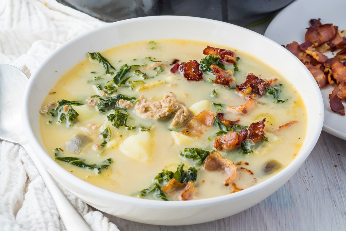 A bowl of creamy whole30 zuppa toscana soup with chopped kale, bacon pieces, potatoes, and sausage in a white ceramic dish.