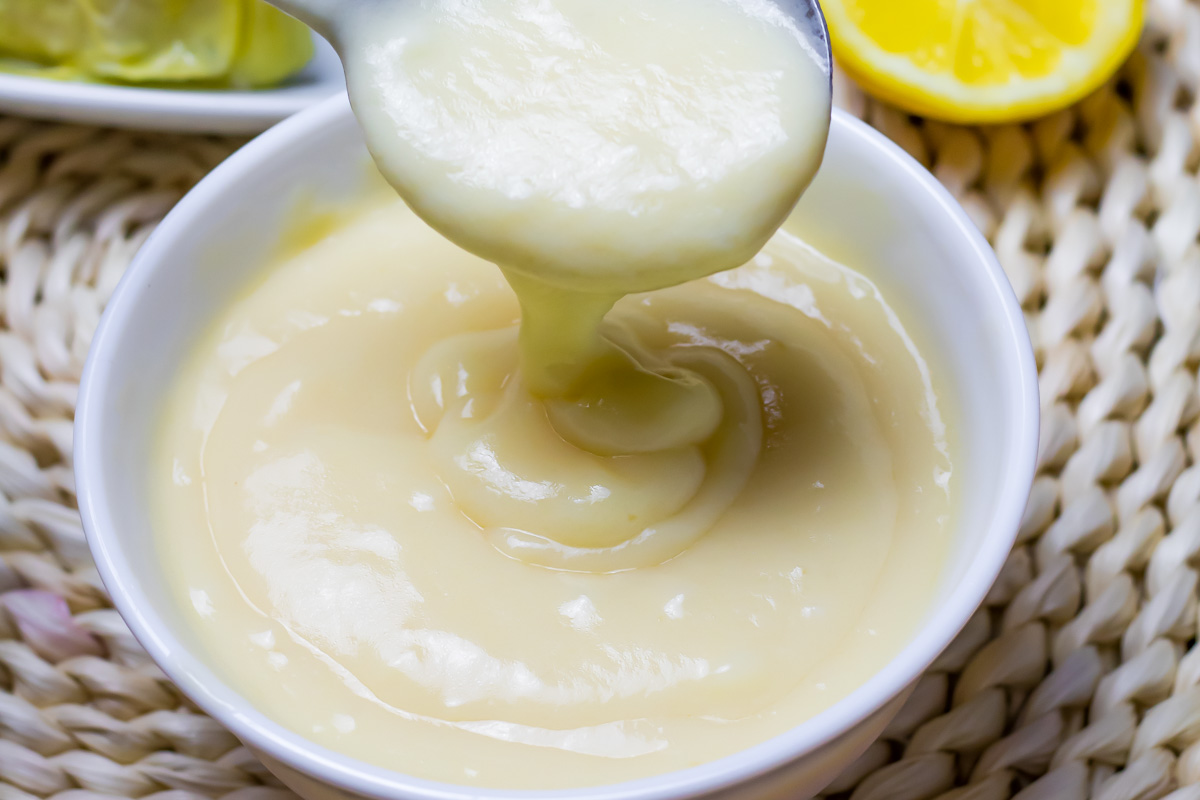 A bowl of creamy, pale yellow avgolemono with a spoon lifting some out; a halved lemon is visible in the background.