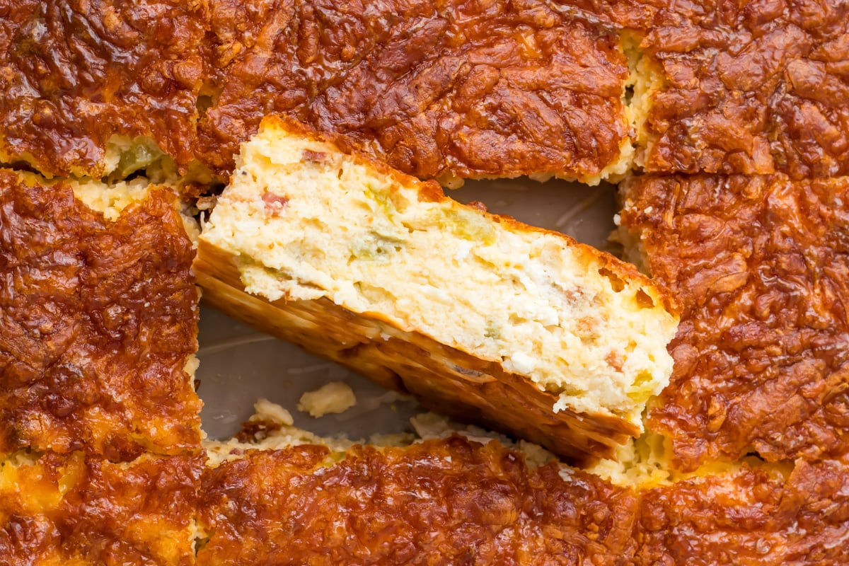 A close-up of a golden-brown cottage cheese egg bake, with one rectangular slice slightly lifted to show its layered, fluffy interior.
