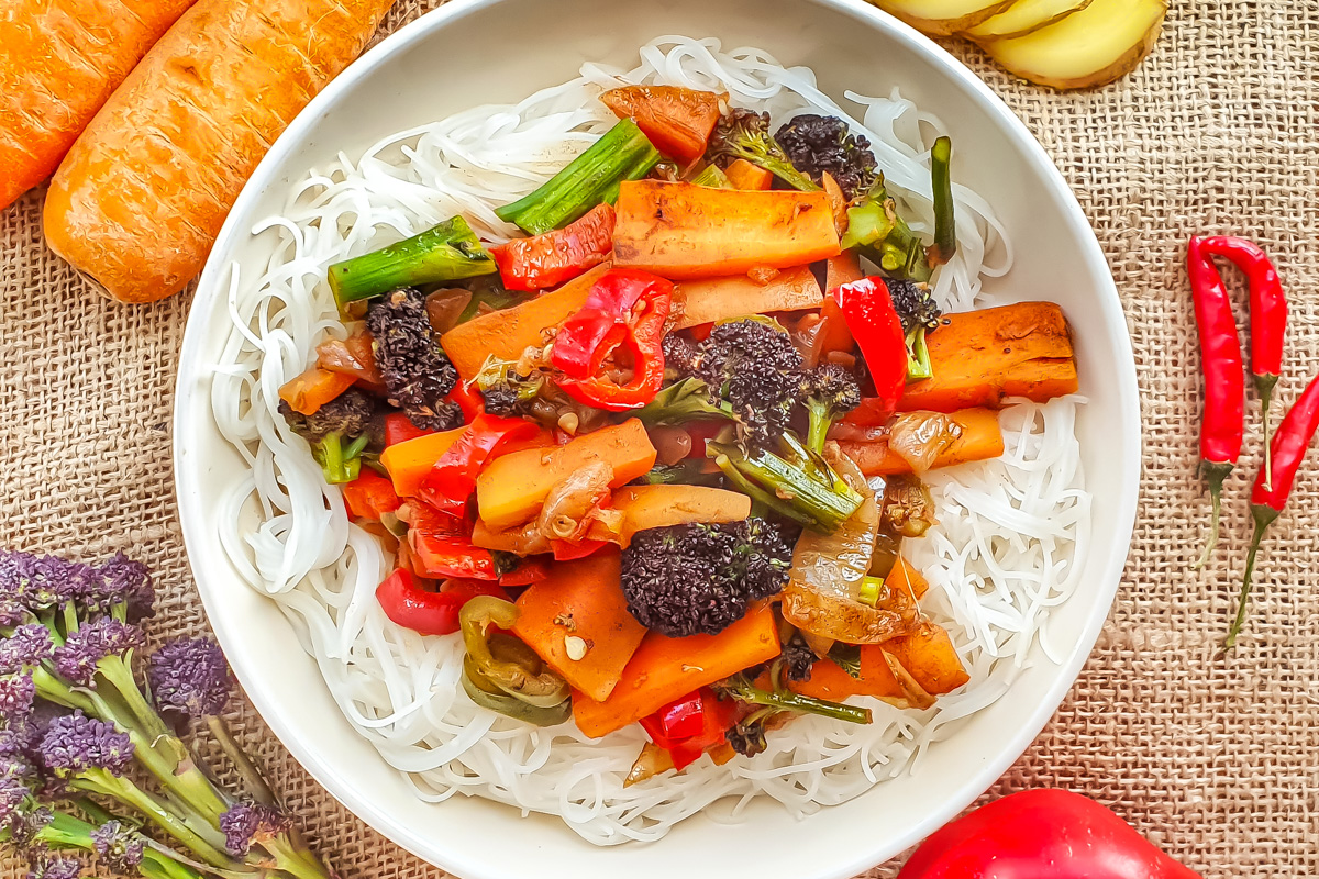 A bowl of whole30 stir-fried vegetables—carrots, bell peppers, broccoli, and green onions—surrounded by fresh ingredients on a burlap surface.