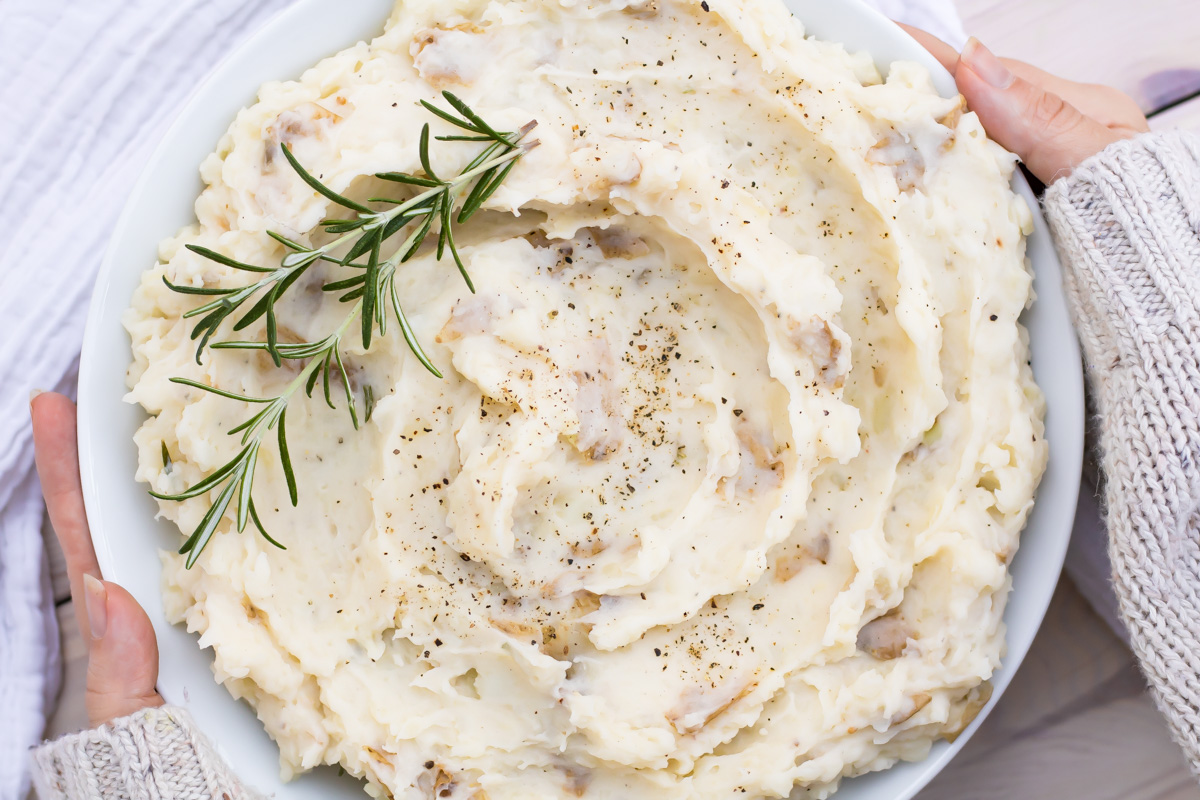 A bowl of whole30 mashed potatoes topped with ground black pepper and garnished with a sprig of rosemary, held by hands in knit sleeves.