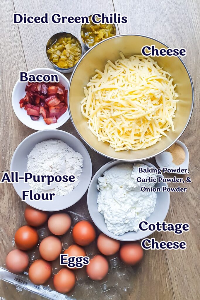 Ingredients for a Cottage Cheese Egg Bake arranged on a wooden surface, featuring diced green chilis, shredded cheese, bacon, flour, cottage cheese, baking powder, spices, and eggs.