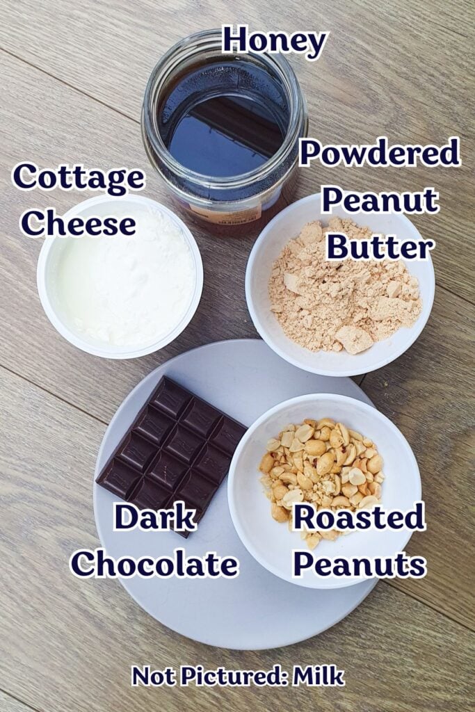 A jar of honey, a bowl of cottage cheese, powdered peanut butter, dark chocolate, and roasted peanuts on a wooden surface; milk not pictured.
