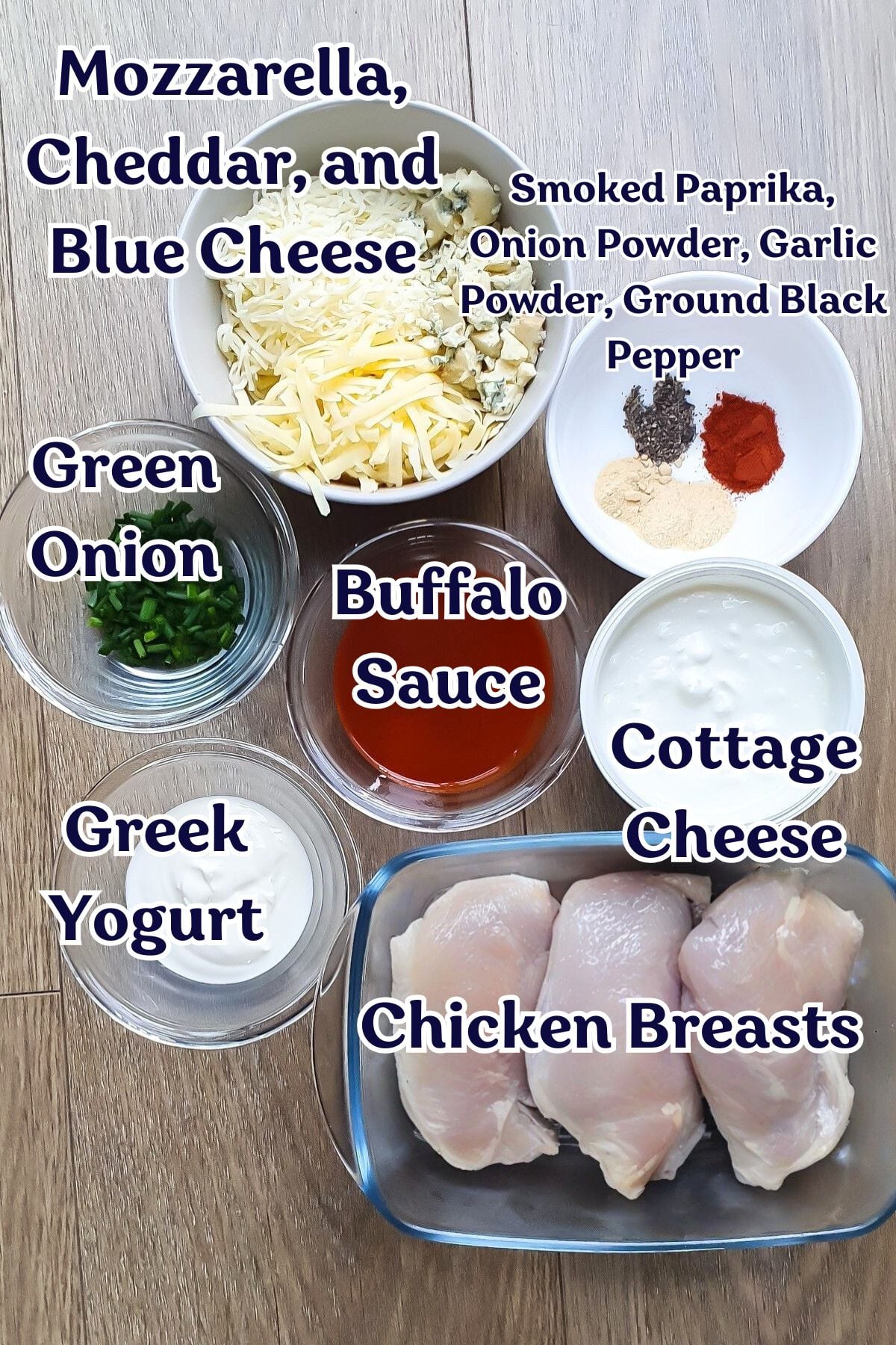 Ingredients for a recipe are displayed in bowls and containers, including cheeses, green onion, Greek yogurt, buffalo sauce, cottage cheese, chicken breasts, and various spices.