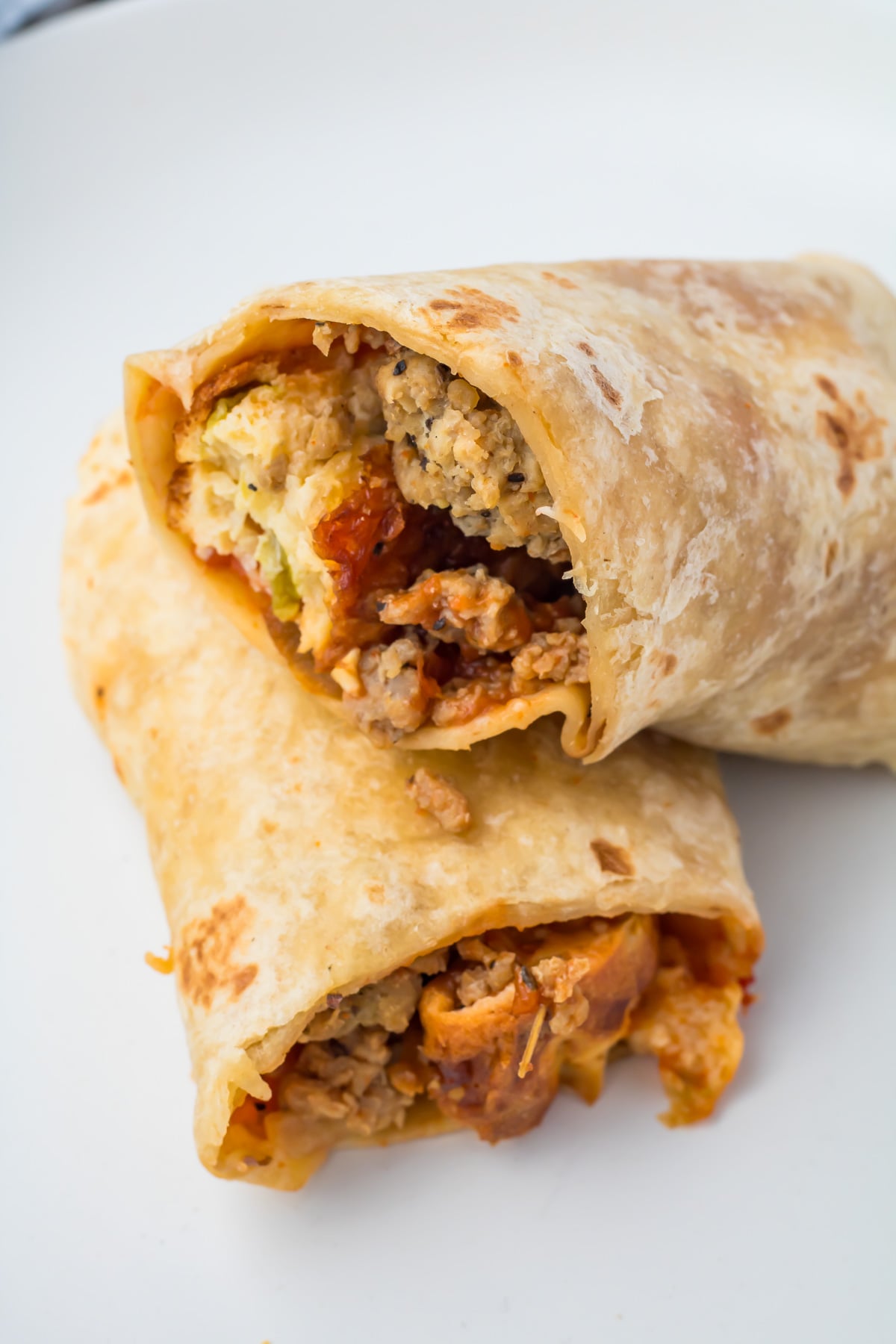 A close-up of two breakfast burritos cut in half, showing a filling of scrambled eggs, sausage, and vegetables on a white plate.