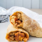 Two halves of a breakfast burrito with eggs, sausage, and cheese are stacked on a white plate, with part of a foil wrapper for meal prep visible in the background.