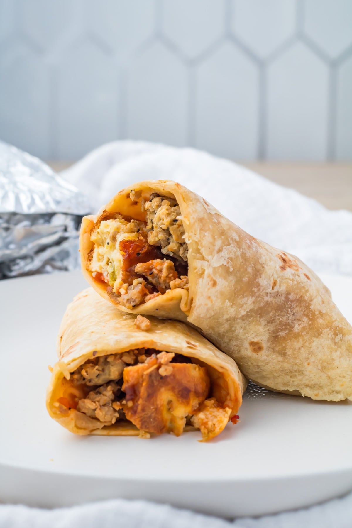 Two halves of a breakfast burrito with eggs, sausage, and cheese are stacked on a white plate, with part of a foil wrapper for meal prep visible in the background.
