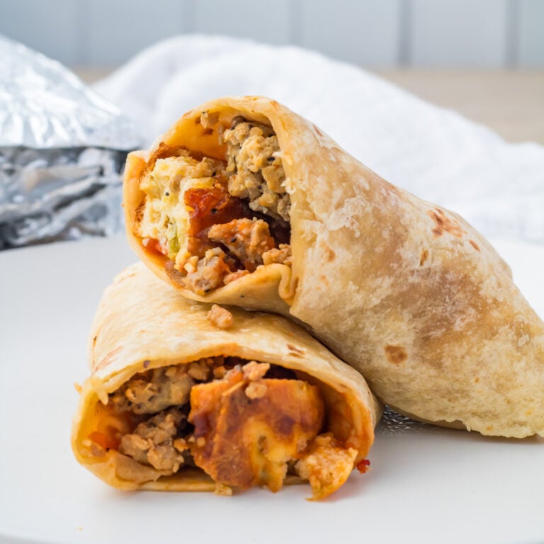 Two breakfast burritos cut in half, filled with scrambled eggs, sausage, and cheese, displayed on a white plate with foil and a white napkin in the background.