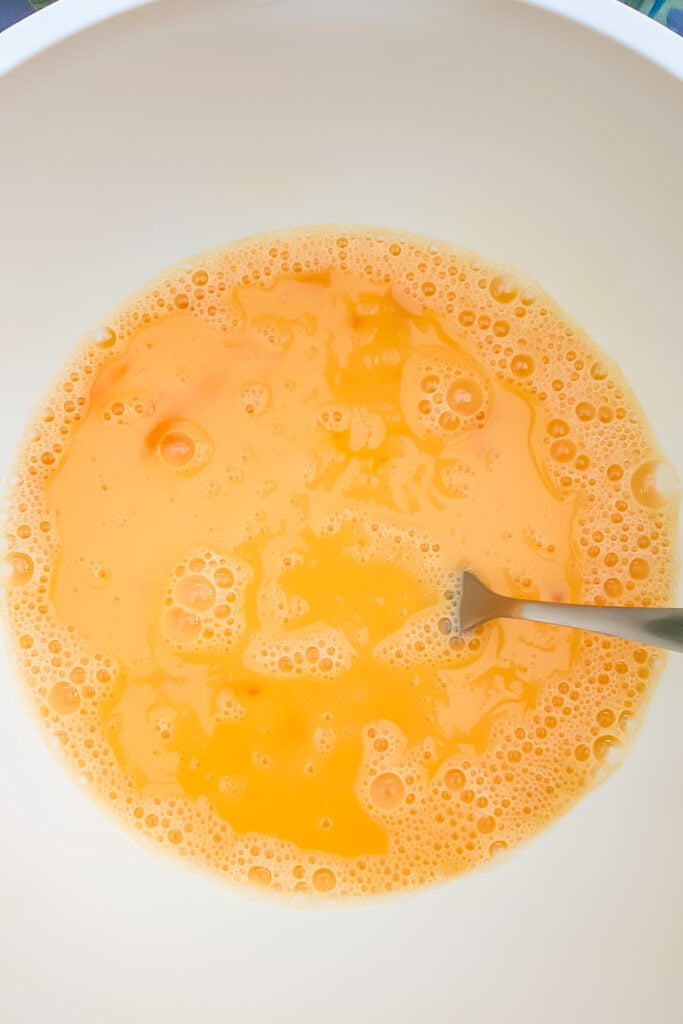 A metal fork in a white bowl with beaten raw eggs, showing a yellow-orange mixture with bubbles&mdash;ready to be used in a delicious Cottage Cheese Egg Bake.