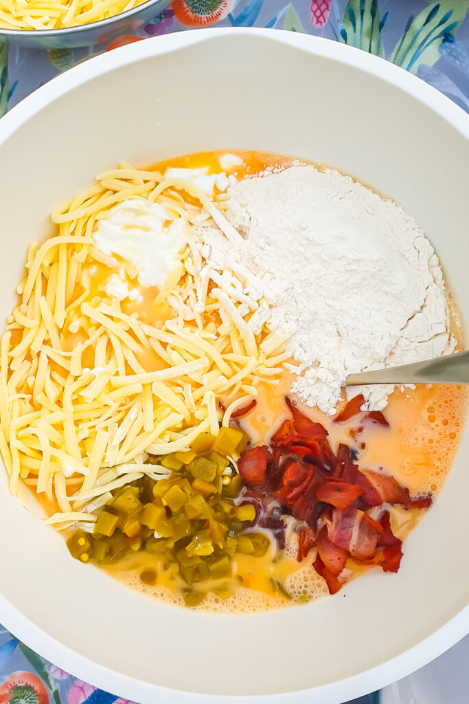A mixing bowl containing shredded cheese, flour, diced jalape&ntilde;os, chopped cooked bacon, beaten eggs, and sour cream, just the right mix for a flavorful Cottage Cheese Egg Bake, with a spoon inside.