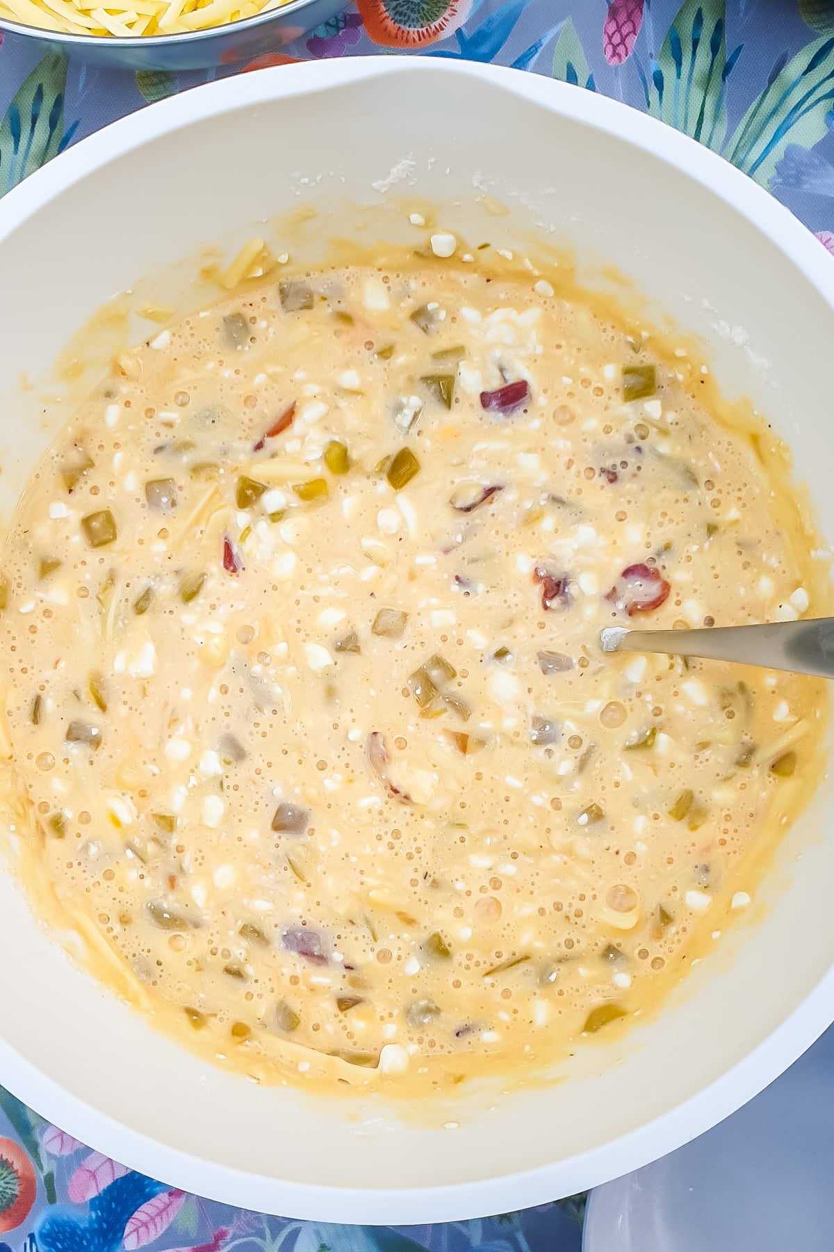 A mixing bowl containing a lumpy, yellowish batter with visible pieces of vegetables and cheese, with a metal spoon resting inside.