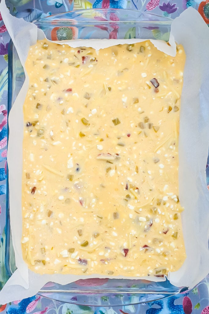 A glass baking dish lined with parchment paper holds an uncooked Cottage Cheese Egg Bake batter, with visible pieces of vegetables and cheese, placed on a colorful, patterned surface.