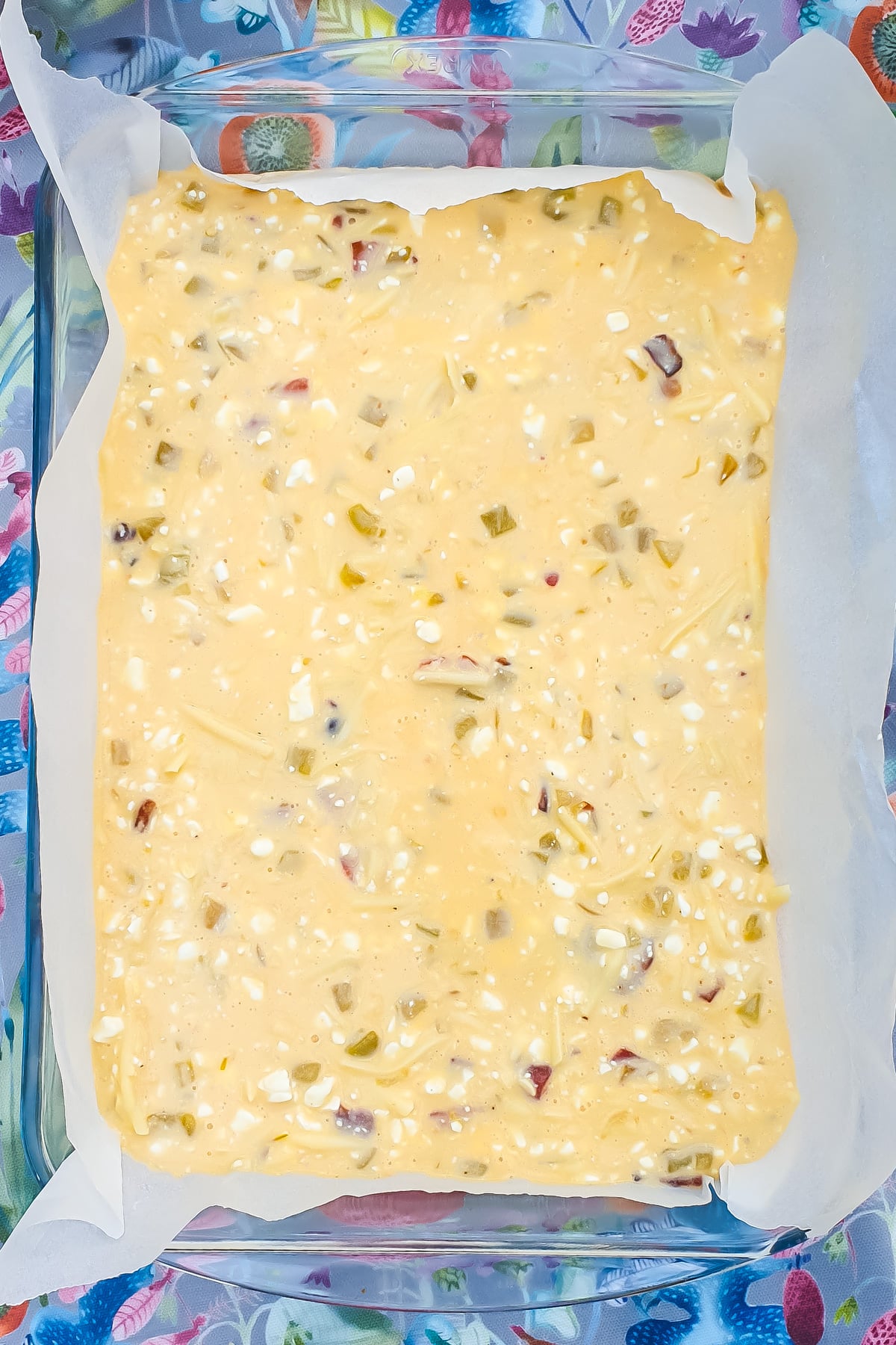 A glass baking dish lined with parchment paper contains an unbaked mixture with visible pieces of vegetables and cheese on a colorful floral surface.