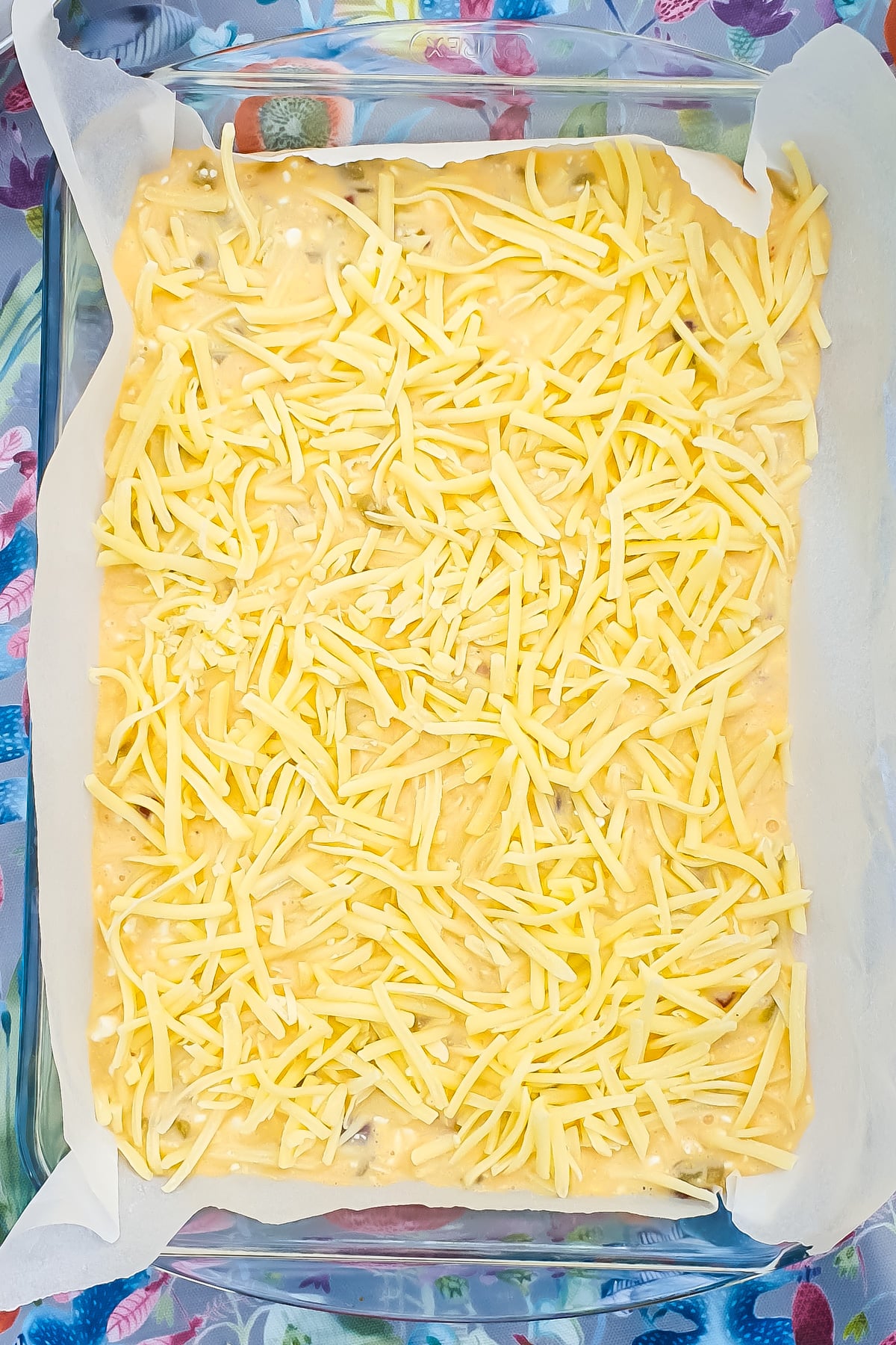 A glass baking dish lined with parchment paper, filled with uncooked batter topped with shredded cheese, on a floral-patterned surface.