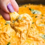A hand with glittery purple nail polish holds a chip dipped in a creamy crockpot buffalo chicken dip with herbs visible in the background.