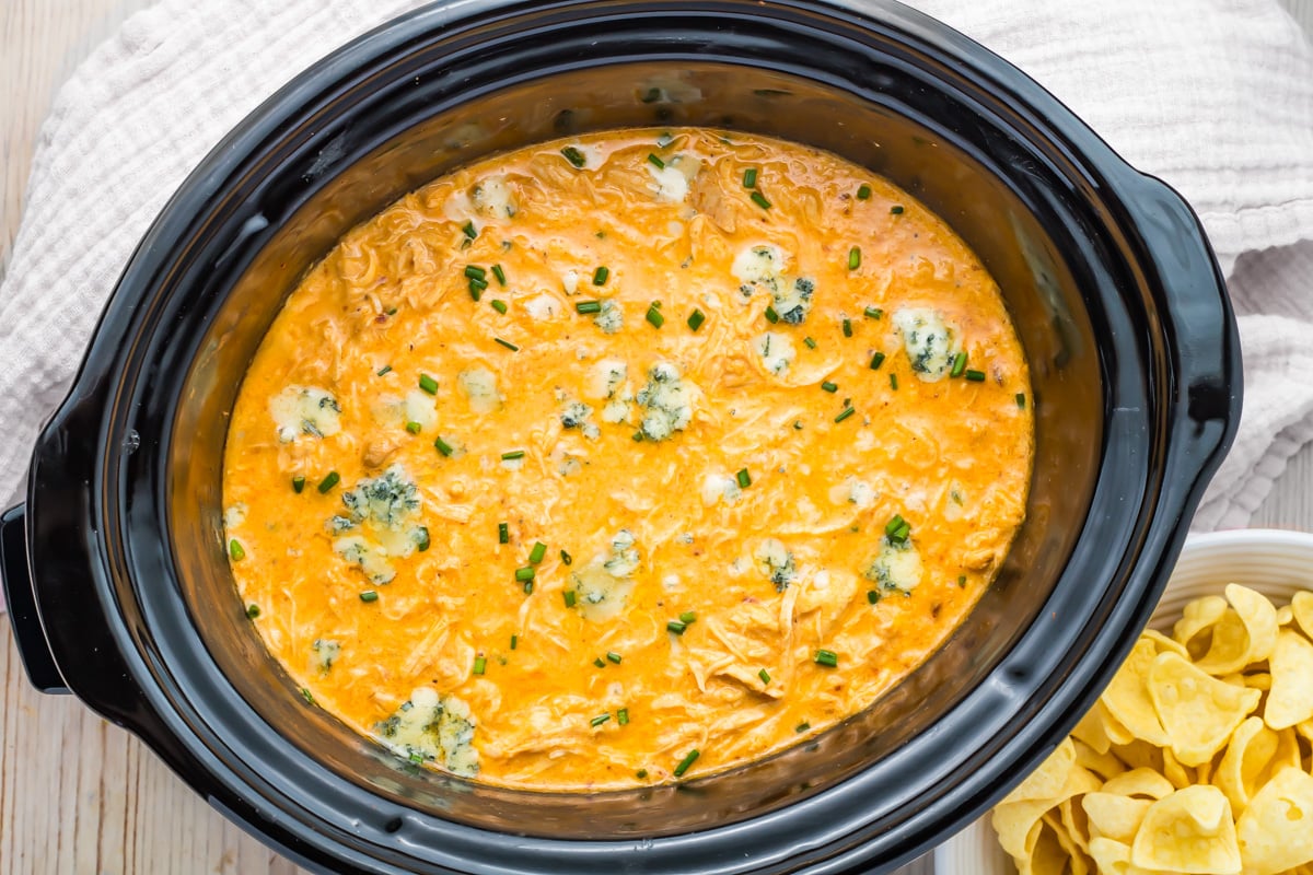 Crockpot Buffalo chicken dip topped with blue cheese and chives in a black slow cooker, with chips in a bowl on the side.