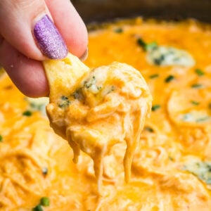 A hand with glittery purple nail polish holds a chip dipped in a creamy crockpot buffalo chicken dip with herbs visible in the background.
