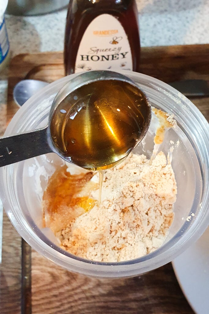 A metal spoon pours honey into a container with a beige powder mixture, with a bottle of honey and kitchen items in the background.
