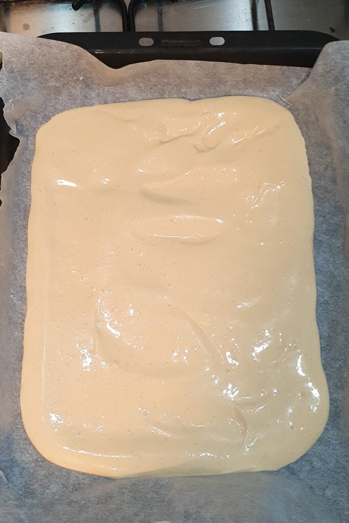 A rectangular baking tray lined with parchment paper holds an even layer of uncooked, frozen cottage cheese bark.