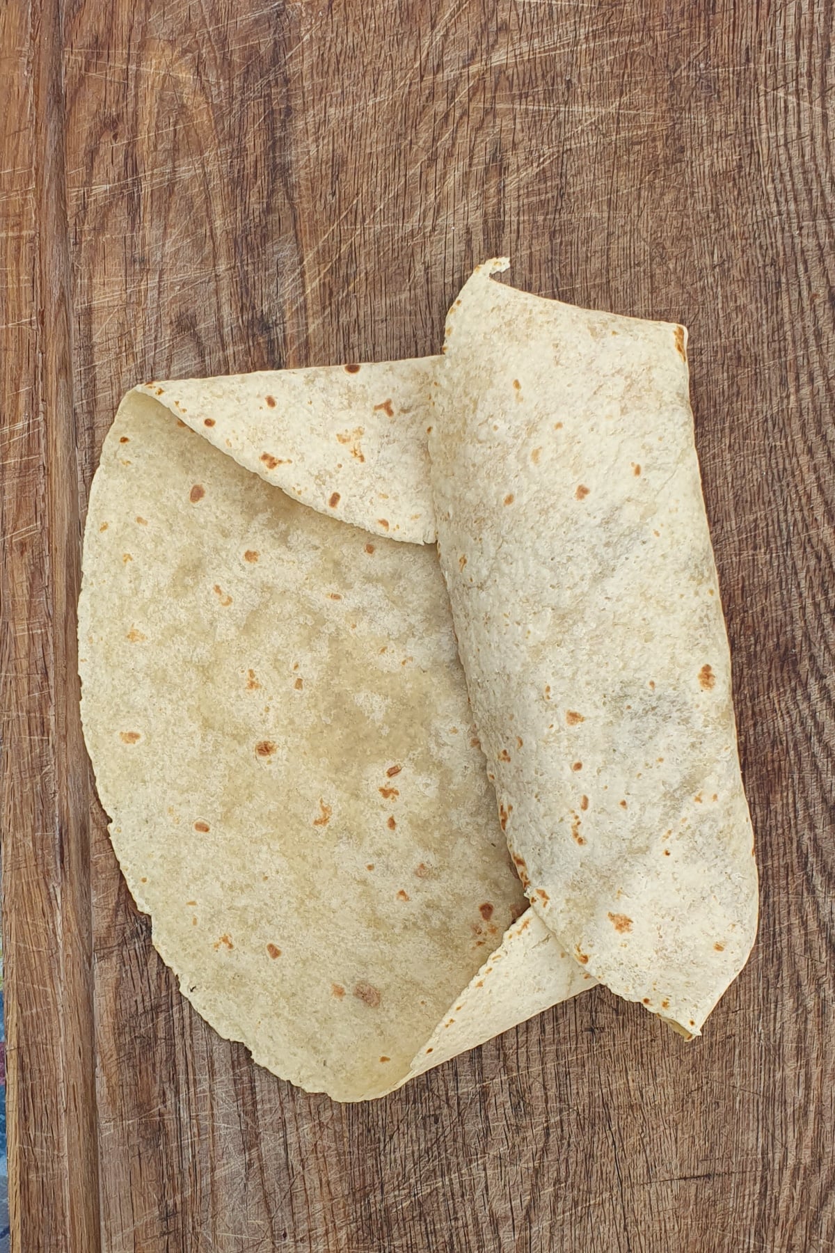 Two flour tortillas are placed on a wooden cutting board, with one tortilla partially folded over the other.