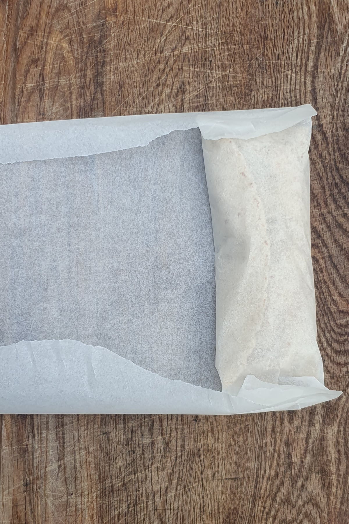 A partially wrapped rectangular food item in parchment paper sits on a wooden surface.