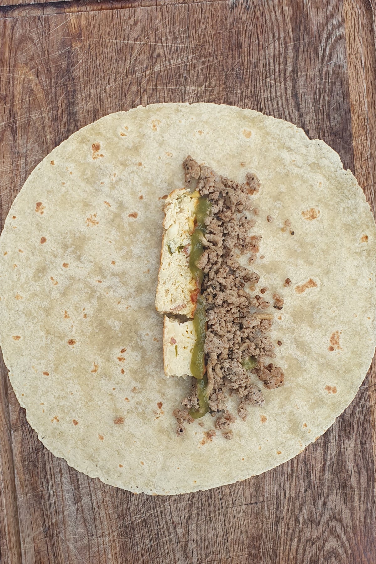 A flour tortilla on a wooden surface with a filling of scrambled eggs, ground meat, and green salsa placed in the center.