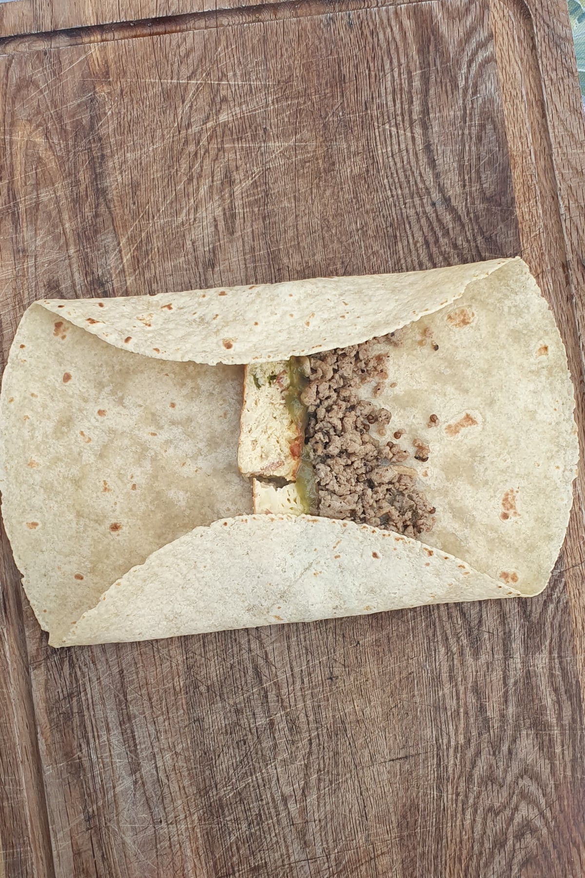 A flour tortilla on a wooden surface, partially folded around seasoned ground meat, tofu, and vegetables.