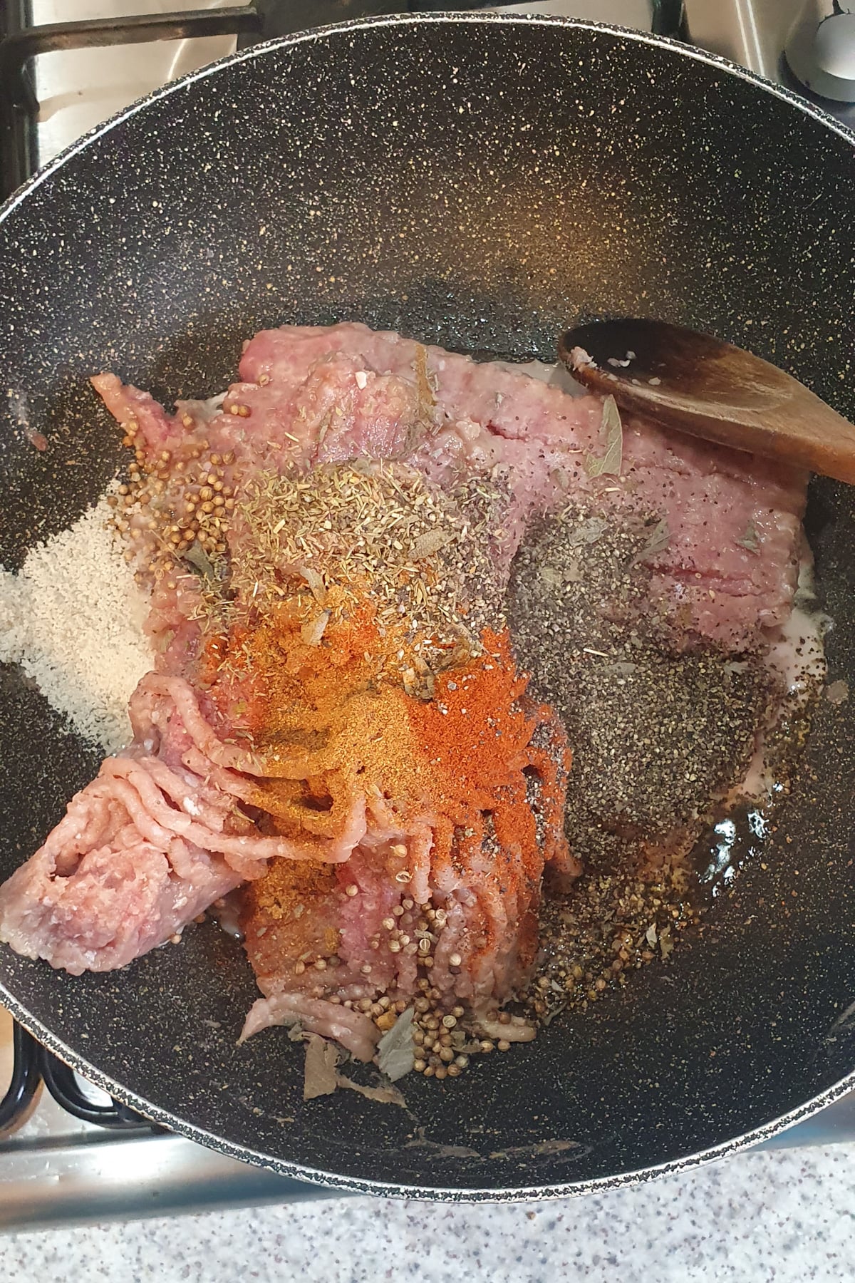 Ground meat in a frying pan with various spices and herbs, including black pepper, paprika, coriander seeds, and bay leaves, next to a wooden spoon on a stovetop.