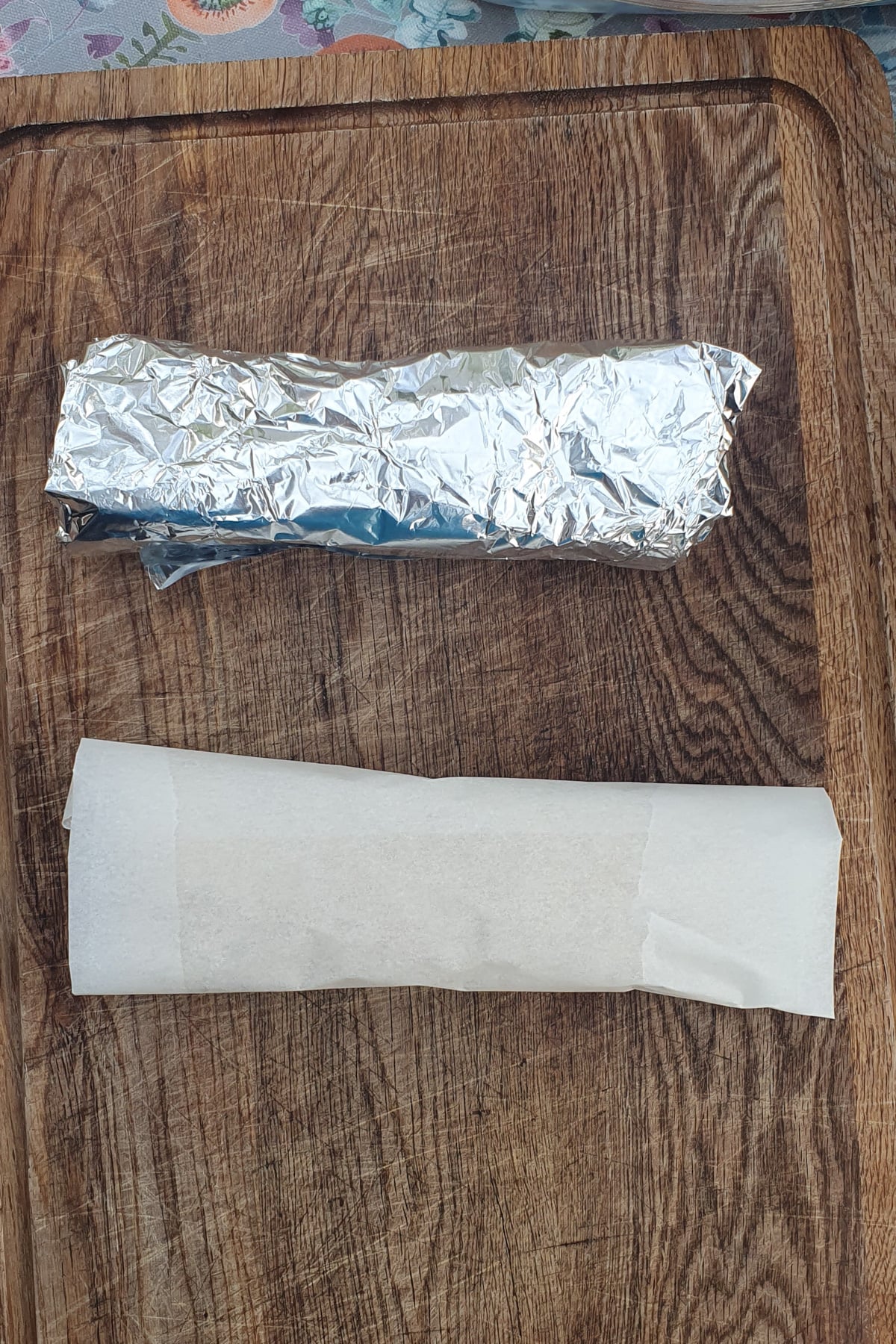 A foil-wrapped roll and a parchment paper-wrapped roll placed side by side on a wooden cutting board.