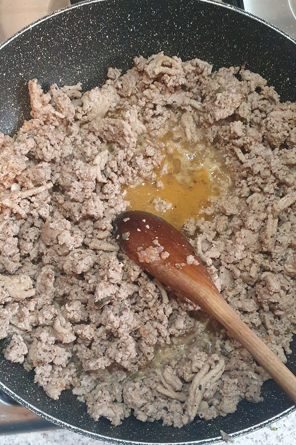 Ground meat being cooked in a frying pan with a wooden spoon resting on top; visible juices pooling in the center.