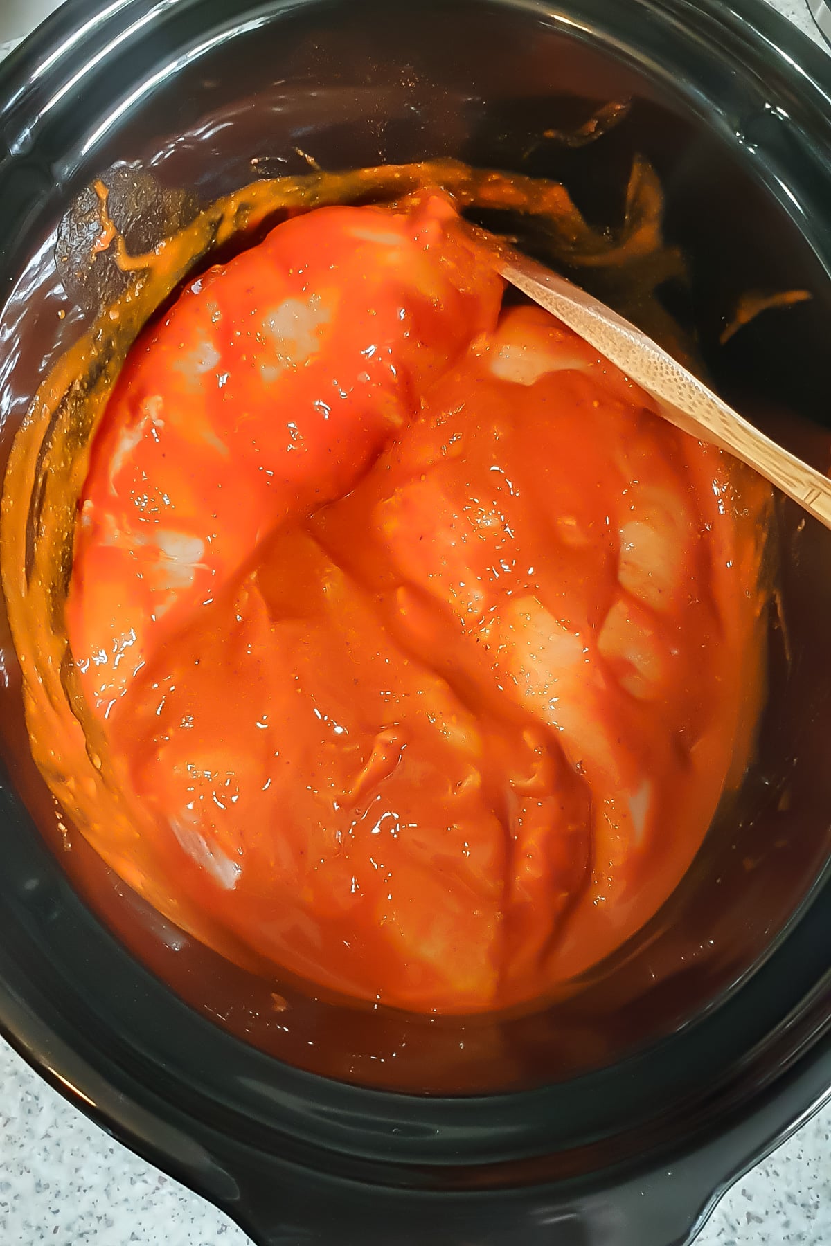 Raw chicken breasts coated in a thick layer of orange-red sauce inside a black slow cooker, with a wooden spoon partially visible.