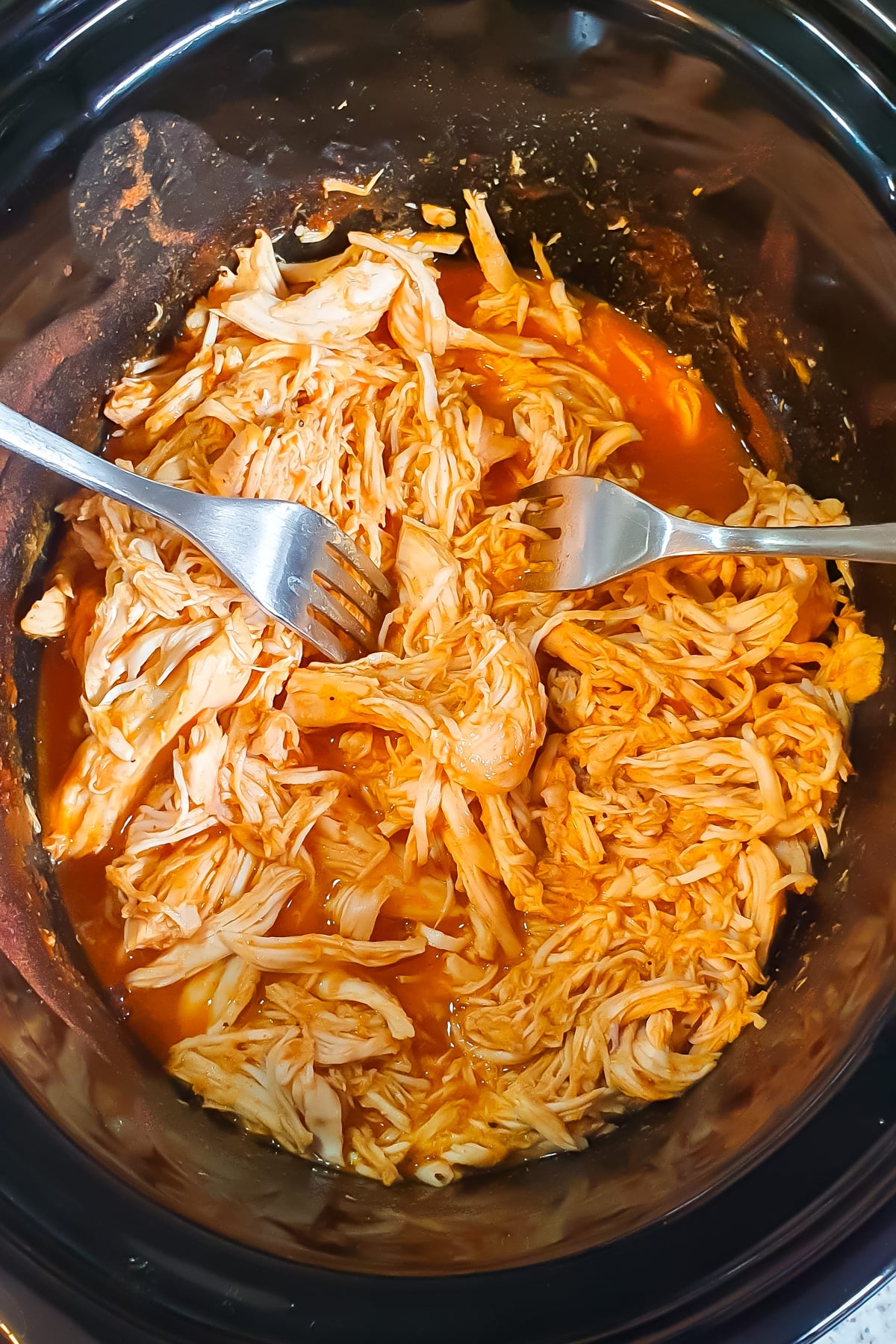 Shredded chicken in a slow cooker with two forks, mixed with orange sauce.