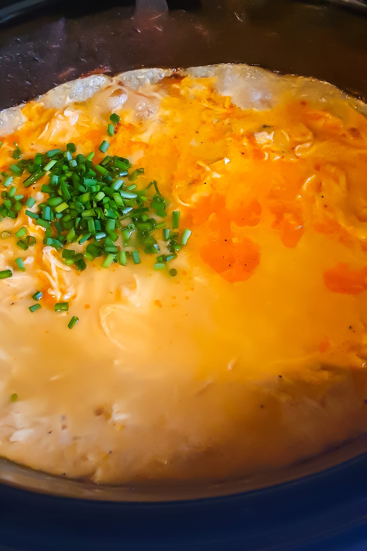 A close-up of creamy buffalo chicken dip in a slow cooker, topped with chopped chives and swirls of orange buffalo sauce.