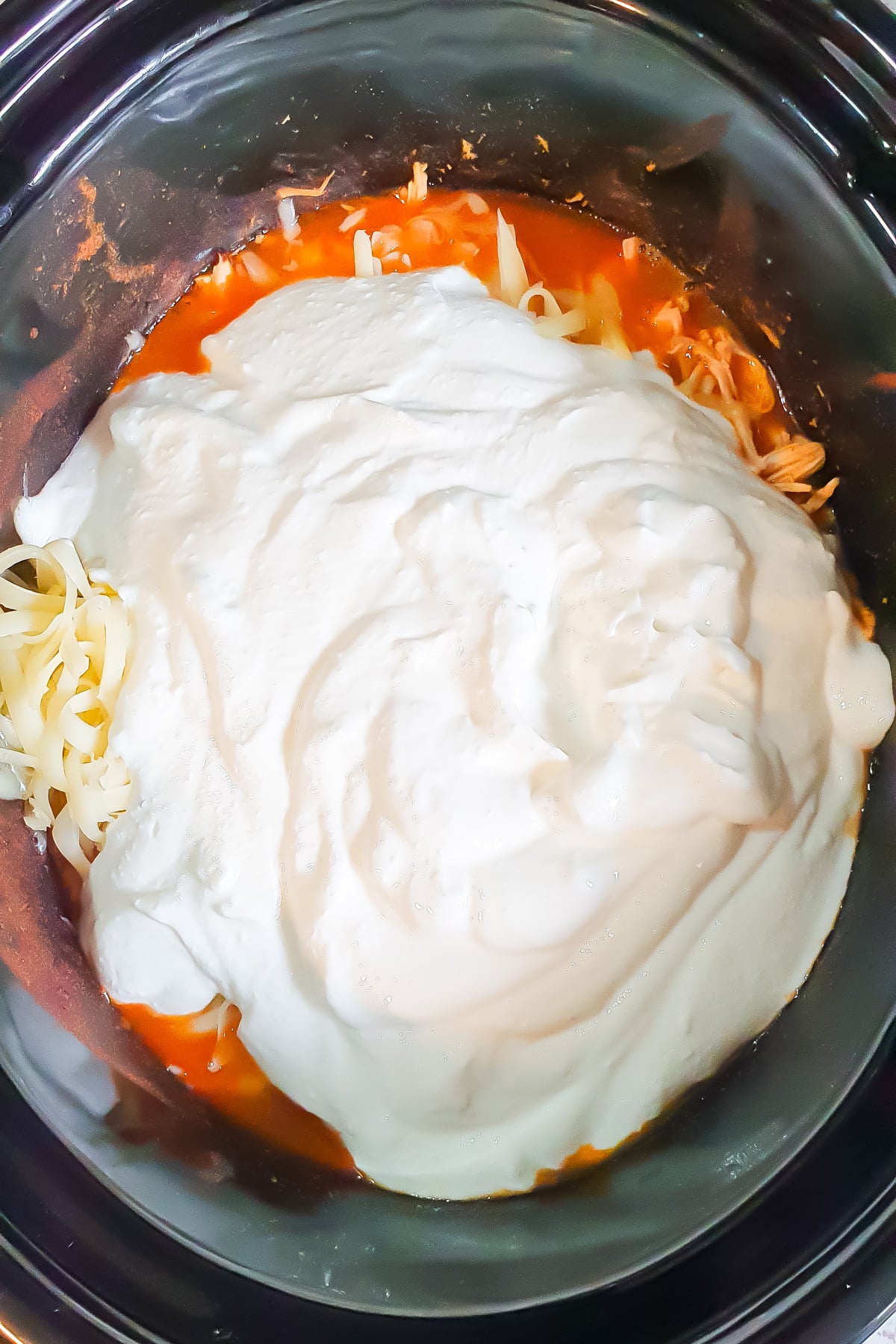Shredded cheese, tomato sauce, and a large amount of sour cream or Greek yogurt are layered inside a slow cooker.
