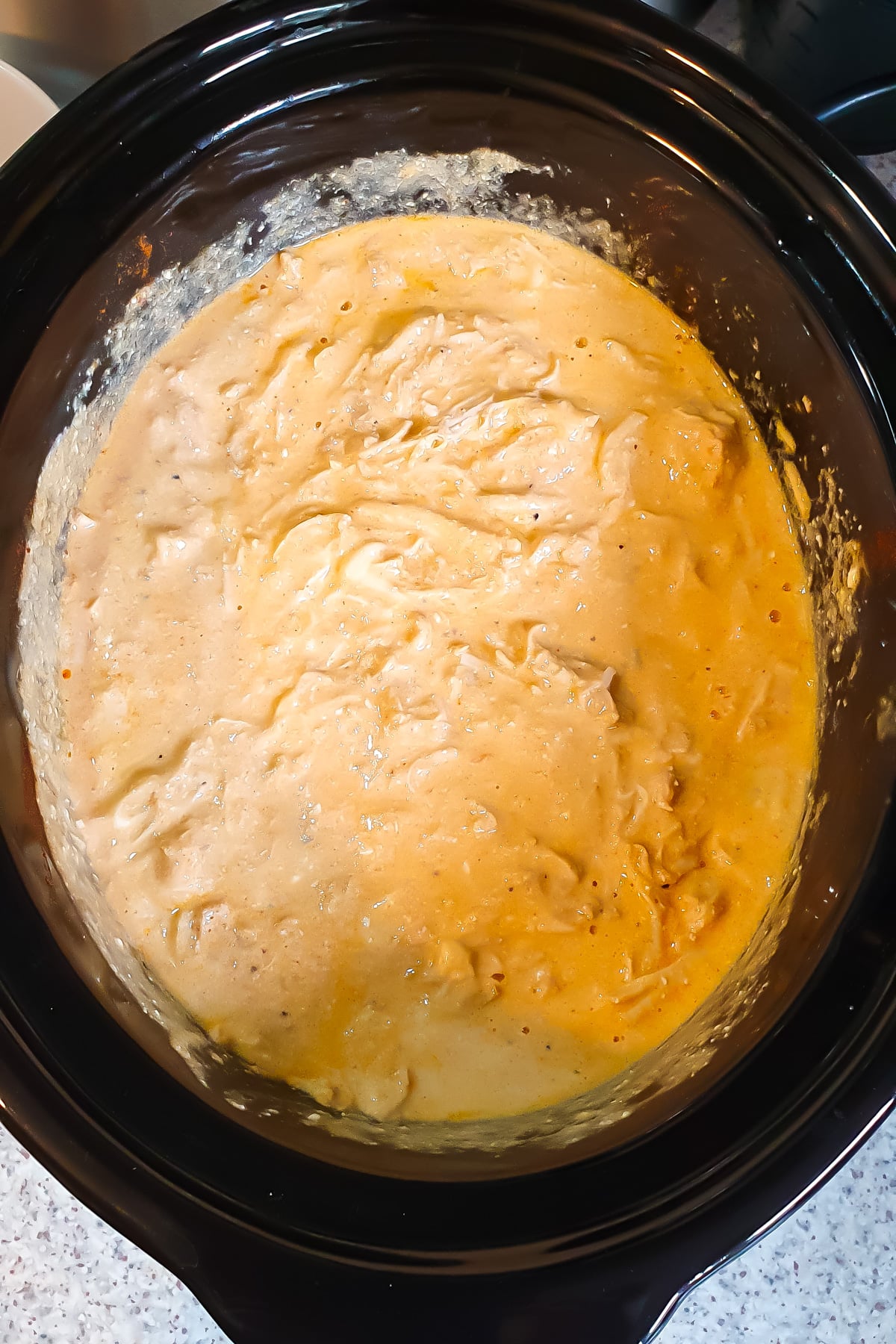 Creamy, orange-colored shredded chicken mixture in a black slow cooker, sitting on a countertop.