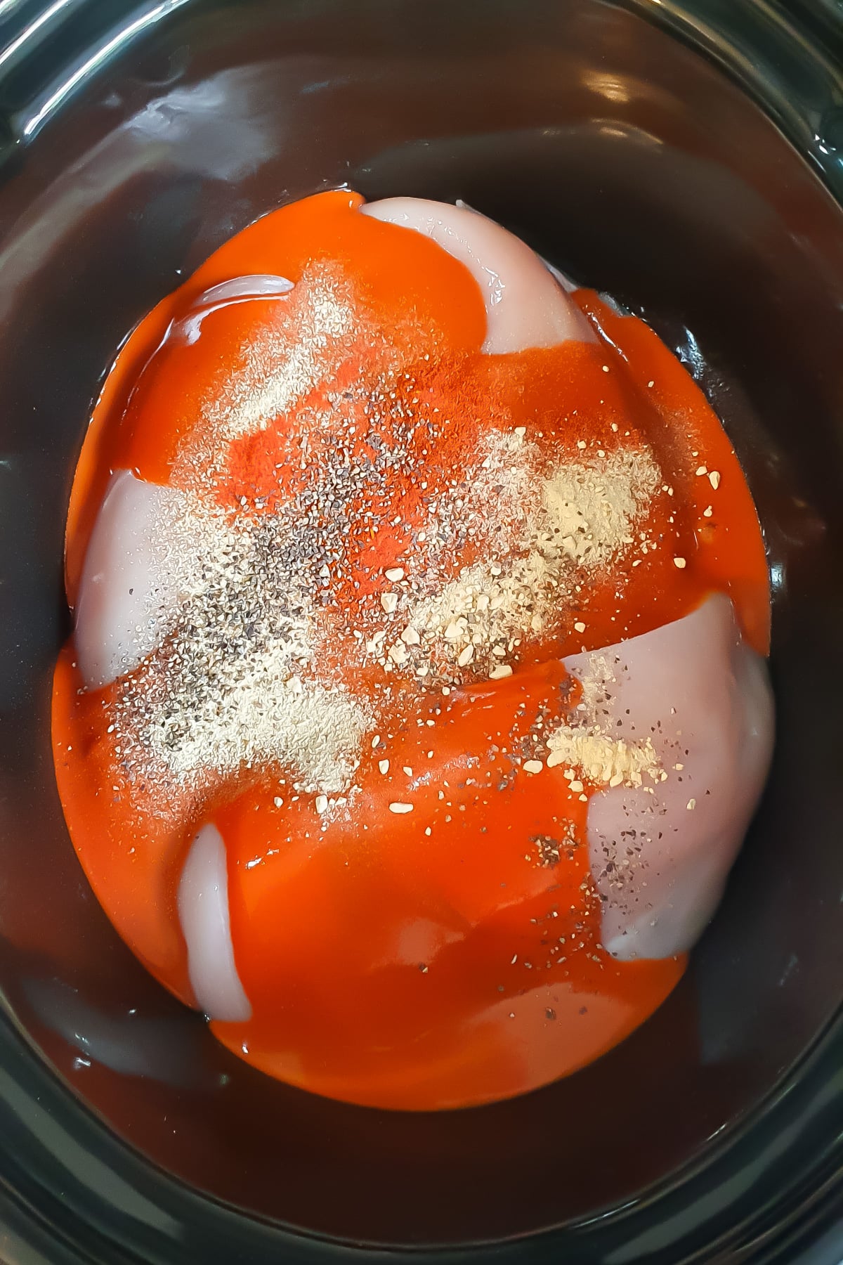 Raw chicken breasts in a slow cooker topped with red sauce and sprinkled with ground black pepper, garlic powder, and other seasonings.
