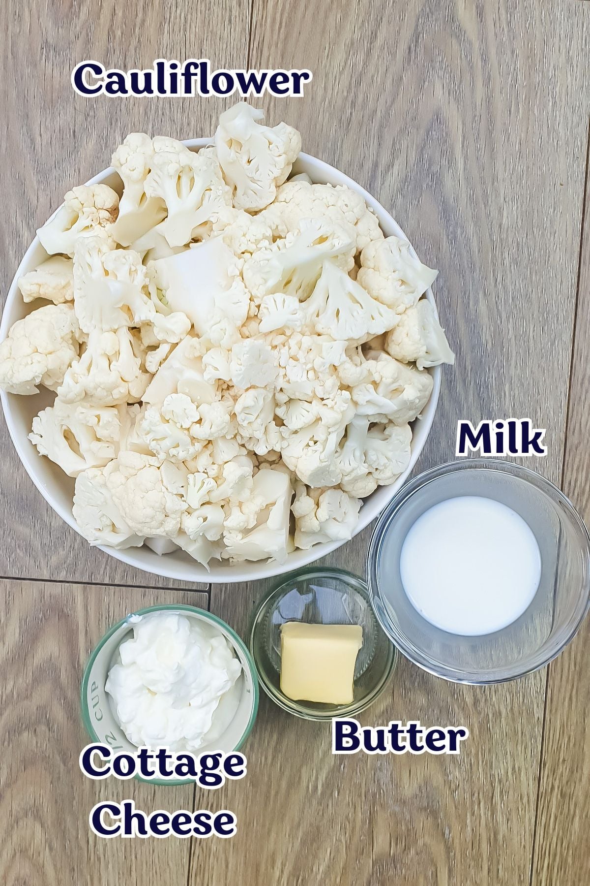 A bowl of cauliflower florets, a small glass of milk, a small bowl of cottage cheese, and a pat of butter are arranged on a wooden surface.