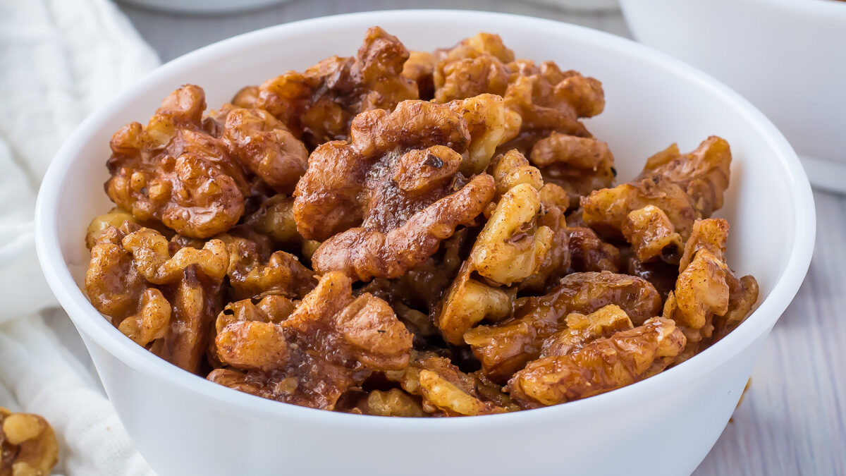 Three white bowls of air fryer candied walnuts, with a cloth and walnuts scattered on a white table.
