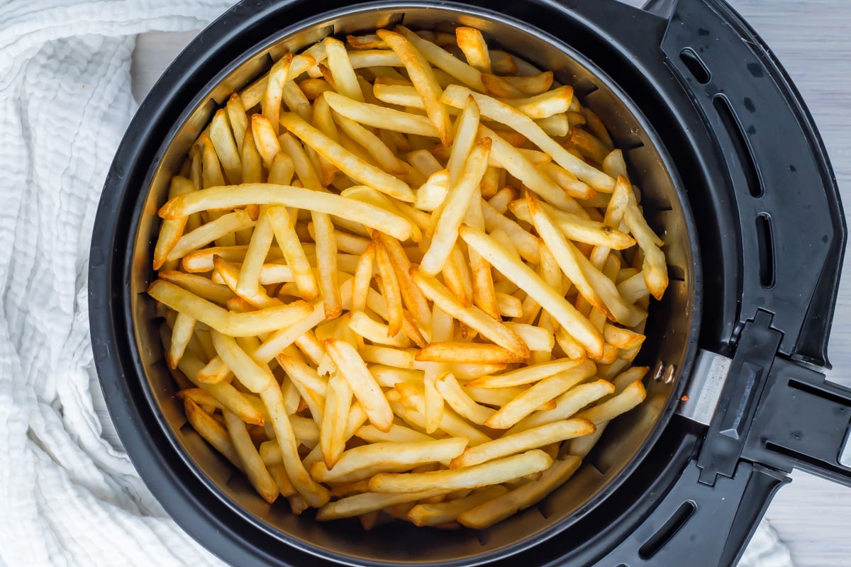 French fries in an air fryer basket, partially cooked with a light golden color, sit next to a white textured cloth.
