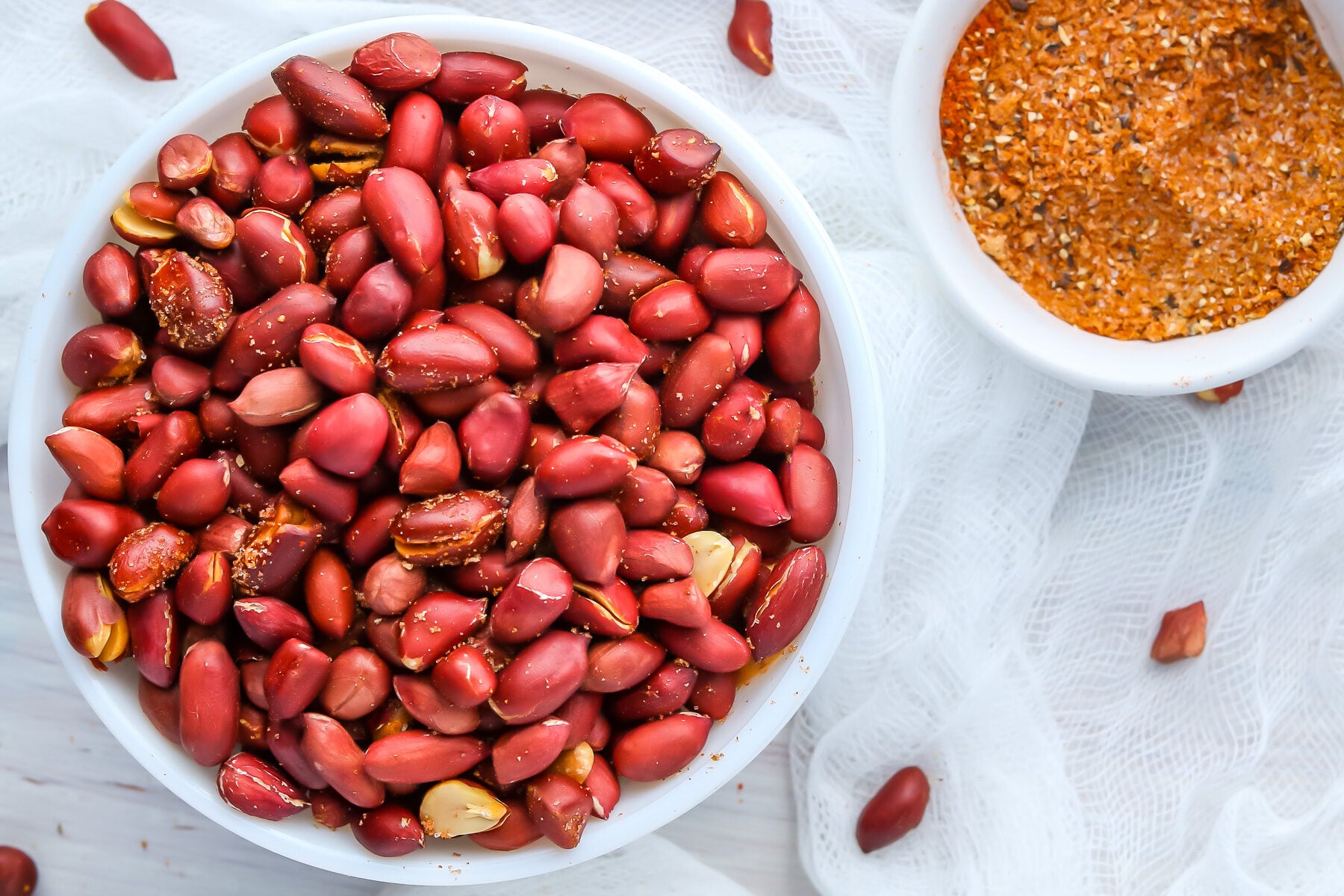 Overhead photo of Air Fryer Peanuts in a white bowl with spice mix in a pinch bowl and peanuts scattered around a white cloth.