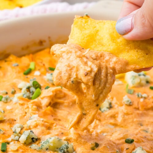 A hand dips a tortilla chip into a creamy, cottage cheese buffalo chicken dip topped with crumbled cheese and chopped green onions.