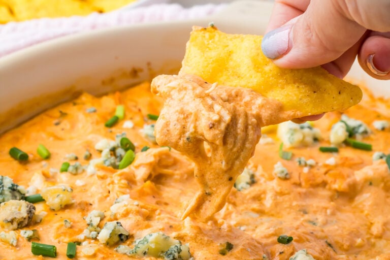 A hand dips a tortilla chip into a creamy, cottage cheese buffalo chicken dip topped with crumbled cheese and chopped green onions.