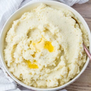 A bowl of cauliflower mash with a pat of melted butter in the center, placed on a light wooden surface with a white cloth nearby.