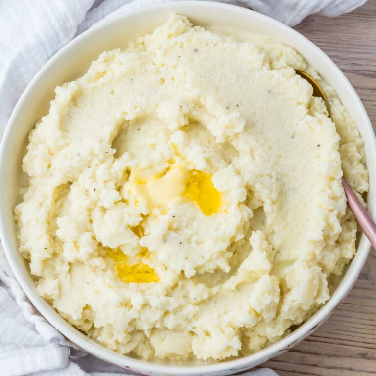 A bowl of cauliflower mash with a pat of melted butter in the center, placed on a light wooden surface with a white cloth nearby.