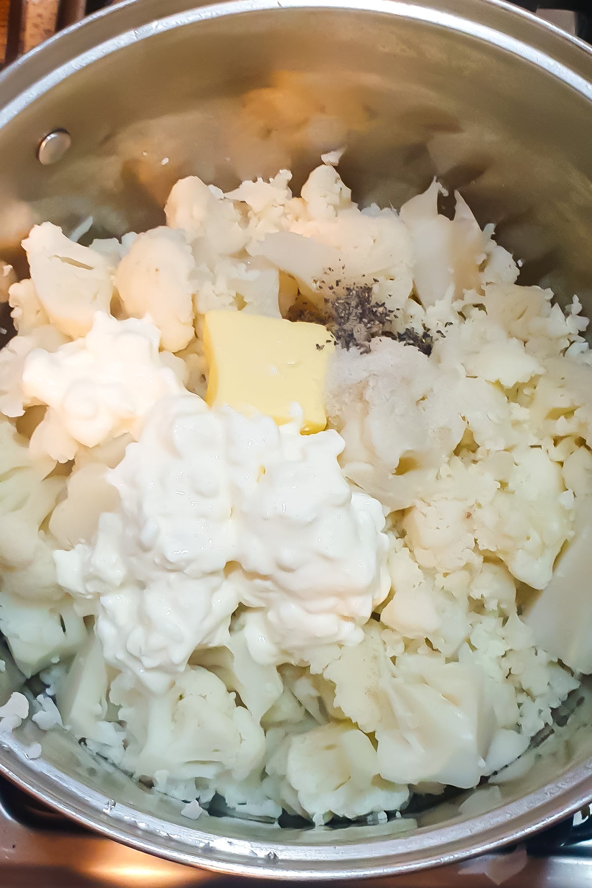A pot filled with cooked cauliflower, butter, cottage cheese, and black pepper, ready to be mixed.