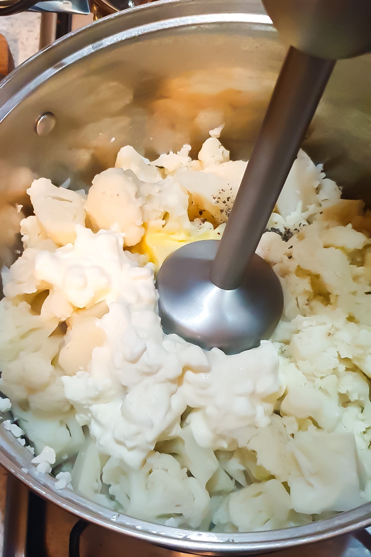 An immersion blender blending cooked cauliflower, butter, and cottage cheese in a pot.