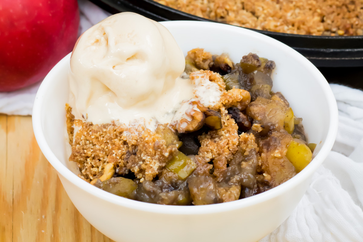 A bowl of keto apple crisp topped with a scoop of vanilla ice cream, with a red apple and baking dish in the background.